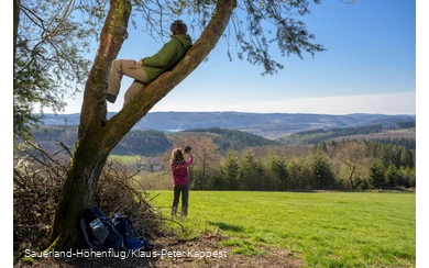 Ein Mann sitzt im Baum und genießt die Aussicht auf den Biggesee. Eine Frau macht im Hintergund ein Selfie am Sauerland-Höhenflug