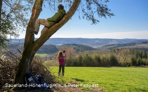 Ein Mann sitzt im Baum und genießt die Aussicht auf den Biggesee. Eine Frau macht im Hintergund ein Selfie am Sauerland-Höhenflug